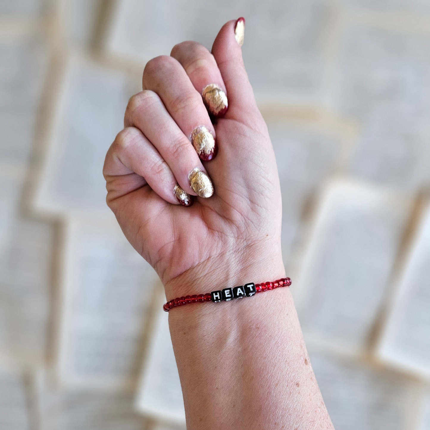 Hand wearing a red and black 'HEAT' bracelet with gold nail polish against a neutral background