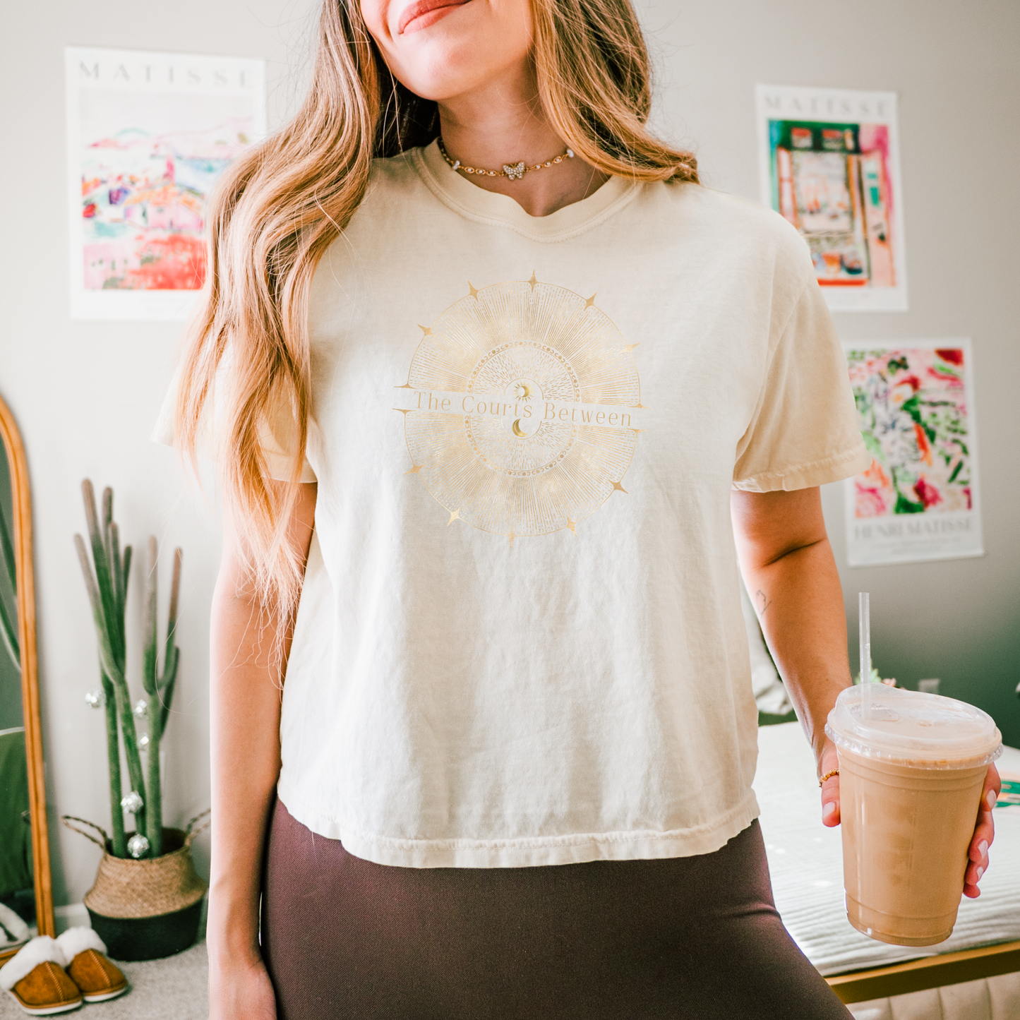 A woman wearing a cream-colored graphic t-shirt with a gold-toned compass design and the text 'The Courts Between'. She is holding an iced coffee in a room decorated with posters and a cactus.