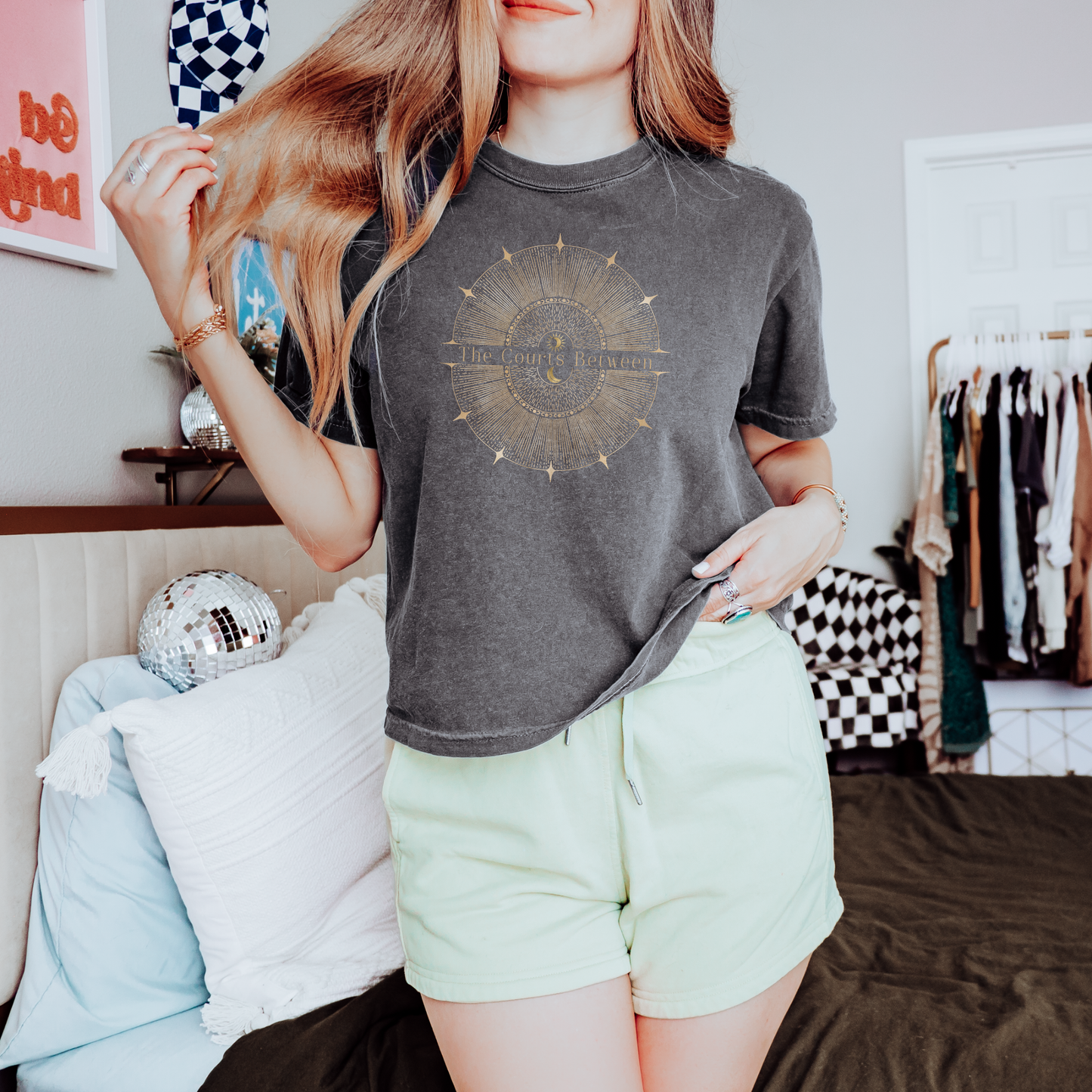 A woman wearing a dark gray washed graphic t-shirt 'The Courts Between' and light green shorts, standing in a bedroom setting.