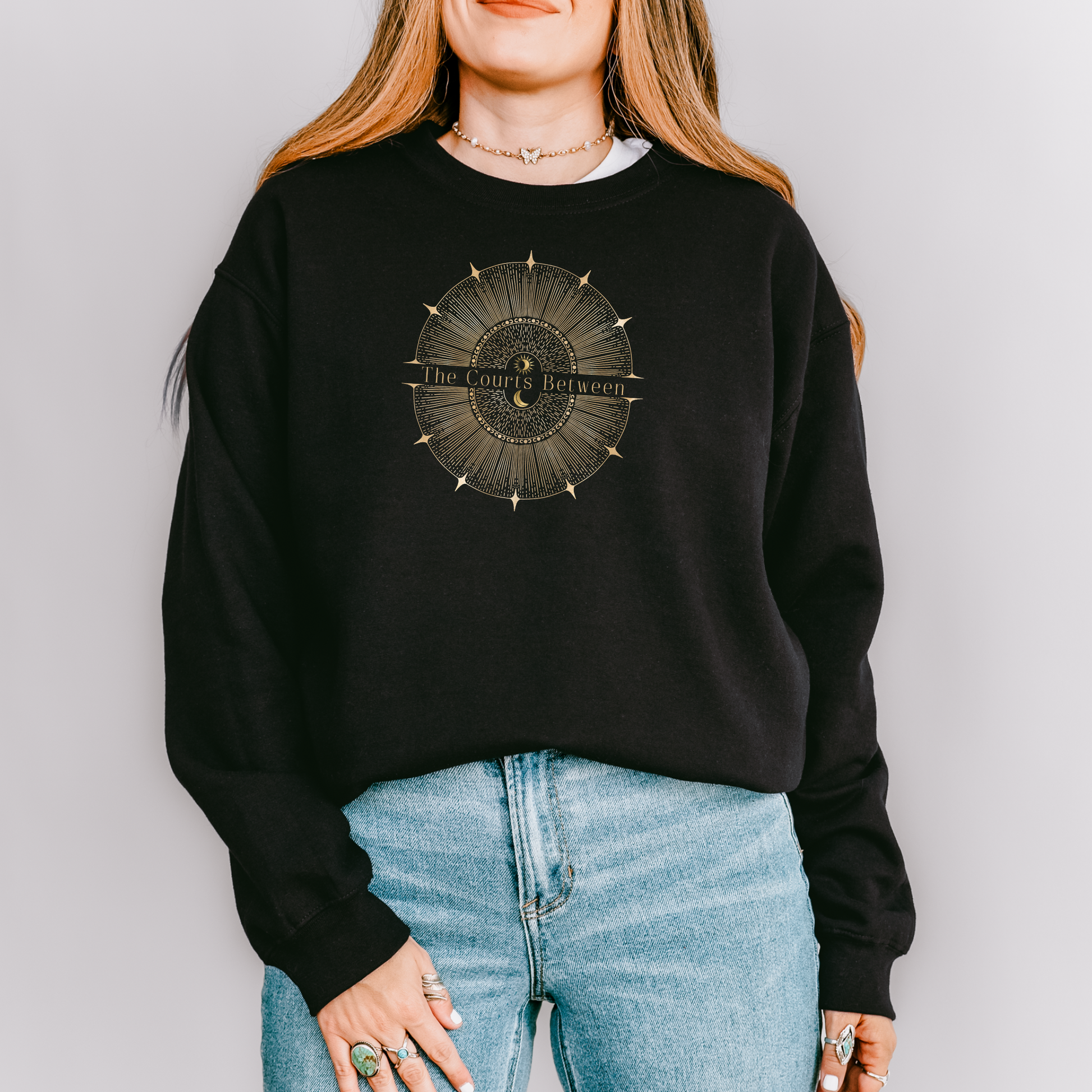 A woman wearing a black crewneck sweatshirt with a circular gold graphic on the chest that reads 'The Courts Between', paired with light blue jeans, against a solid light gray background.