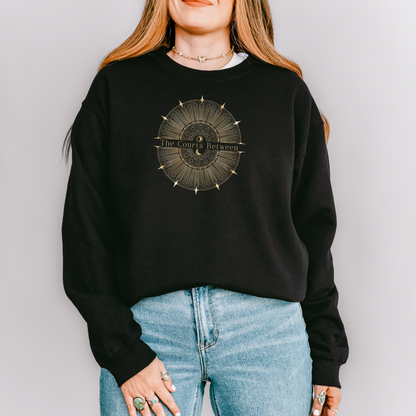 A woman wearing a black crewneck sweatshirt with a circular gold graphic on the chest that reads 'The Courts Between', paired with light blue jeans, against a solid light gray background.