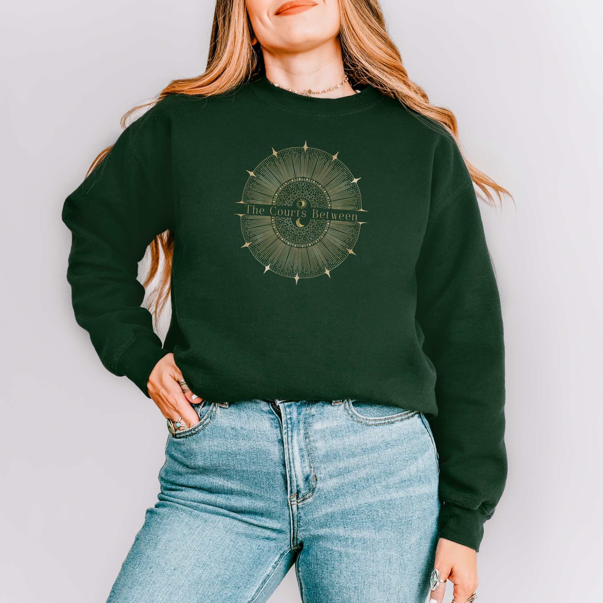 A woman wearing a dark forest green crewneck sweatshirt with a circular gold-toned graphic on the chest that reads 'The Courts Between', paired with light blue jeans, against a solid light gray background.