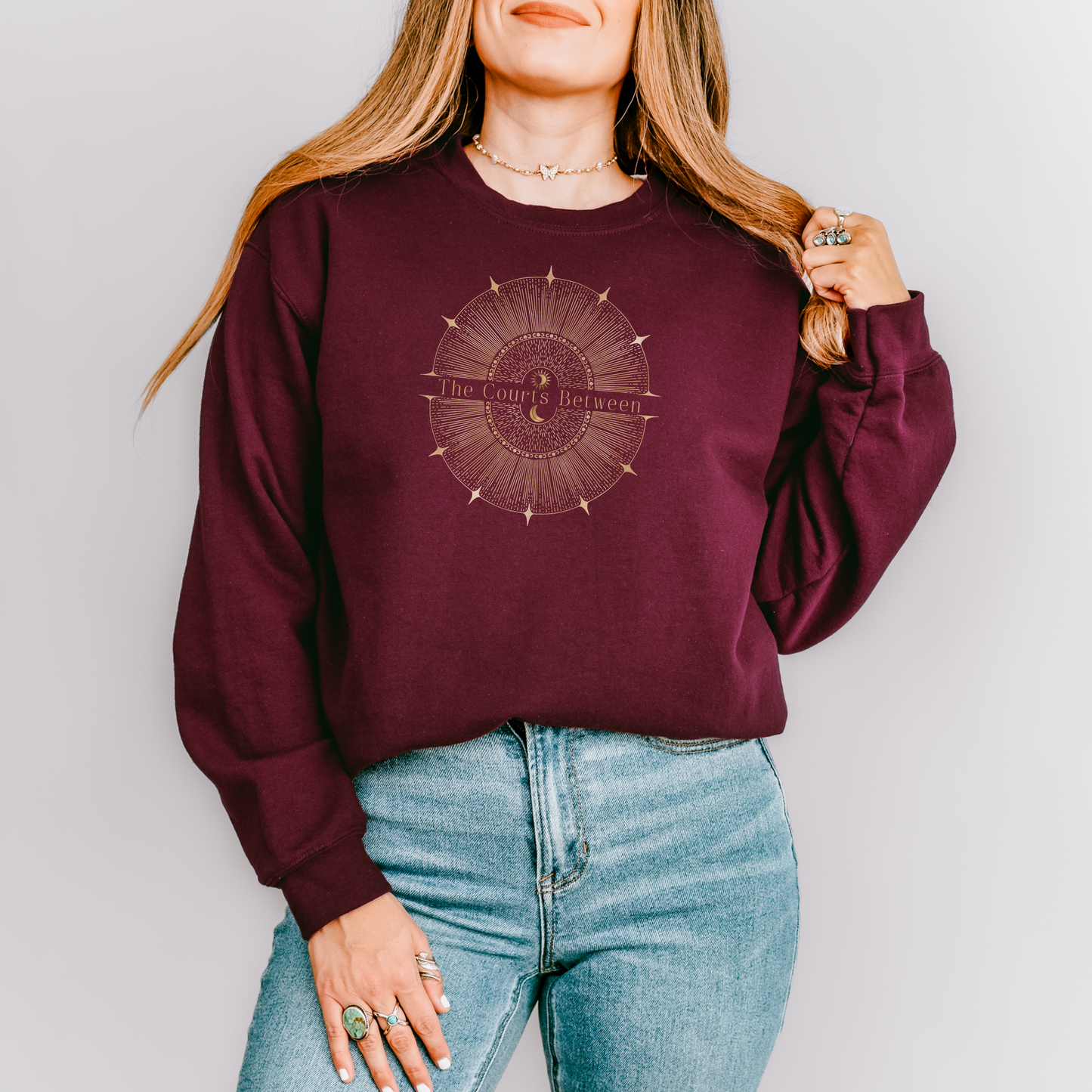 A woman wearing a maroon crewneck sweatshirt with a circular celestial graphic that reads 'The Courts Between', and light blue jeans, standing against a solid light gray background.