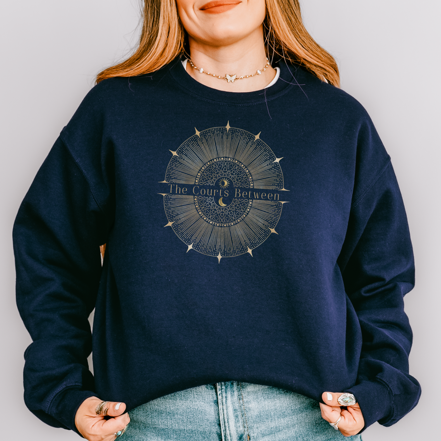 A woman wearing a navy blue crewneck sweatshirt with a circular celestial graphic on the chest that reads 'The Courts Between', paired with light blue jeans, against a plain light gray background.