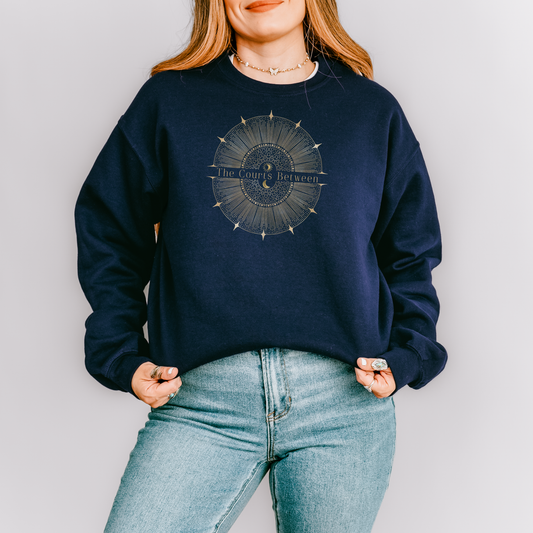A woman wearing a navy blue crewneck sweatshirt with a circular celestial graphic on the chest that reads 'The Courts Between', paired with light blue jeans, against a plain light gray background.