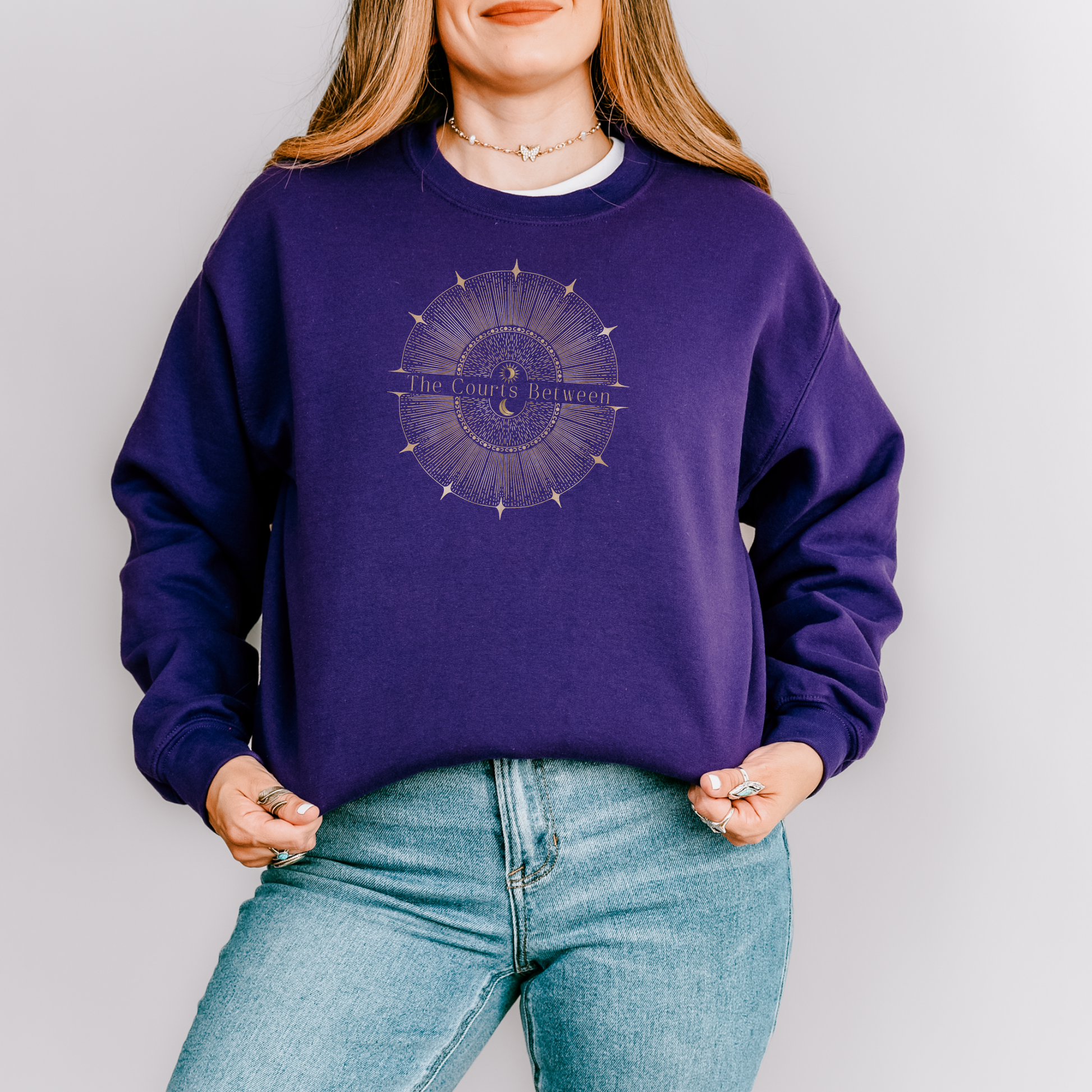 A woman wearing a purple crewneck sweatshirt with a circular graphic on the chest that reads 'The Courts Between', paired with light blue jeans, against a plain light gray background.