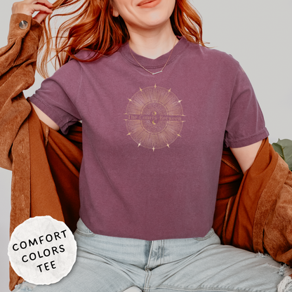 A woman wearing a mauve-purple Comfort Colors graphic t-shirt with a celestial design and the words "The Courts Between". is printed across the center of the design, a brown cardigan and light-wash jeans.
