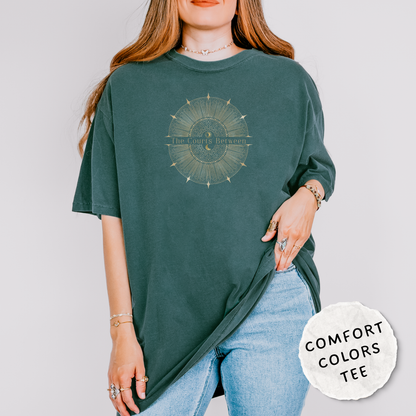 A woman wearing an oversized dark teal-green t-shirt with a celestial design and the words "The Courts Between". is printed across the center of the design. She is wearing light-wash blue jeans and jewelry, standing against a light gray studio background.