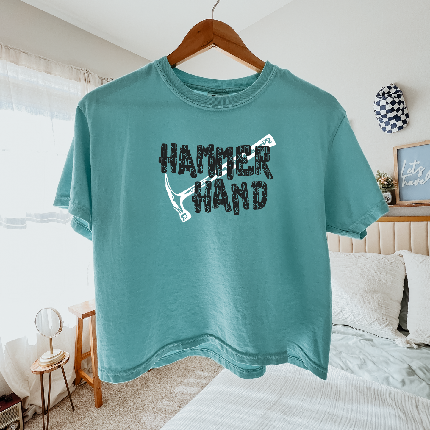 Teal t-shirt with 'HAMMER HAND' text on a hanger in a bedroom.