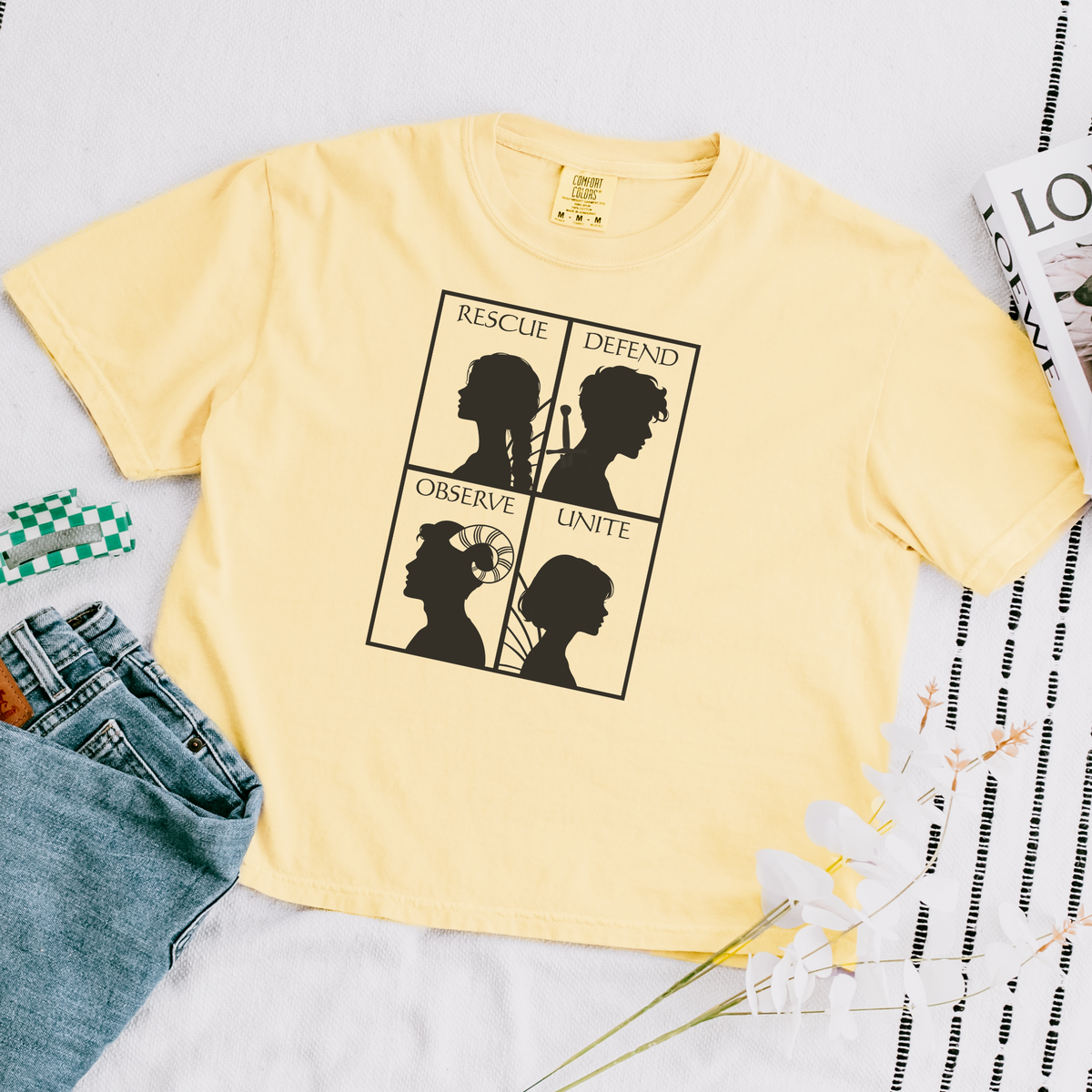 A pale yellow short-sleeve t-shirt laid flat on a white surface, featuring a black graphic of four silhouettes in a grid with the words RESCUE, DEFEND, OBSERVE, and UNITE. Blue jeans, a green-and-white checkered cloth, and printed paper props are placed around it.