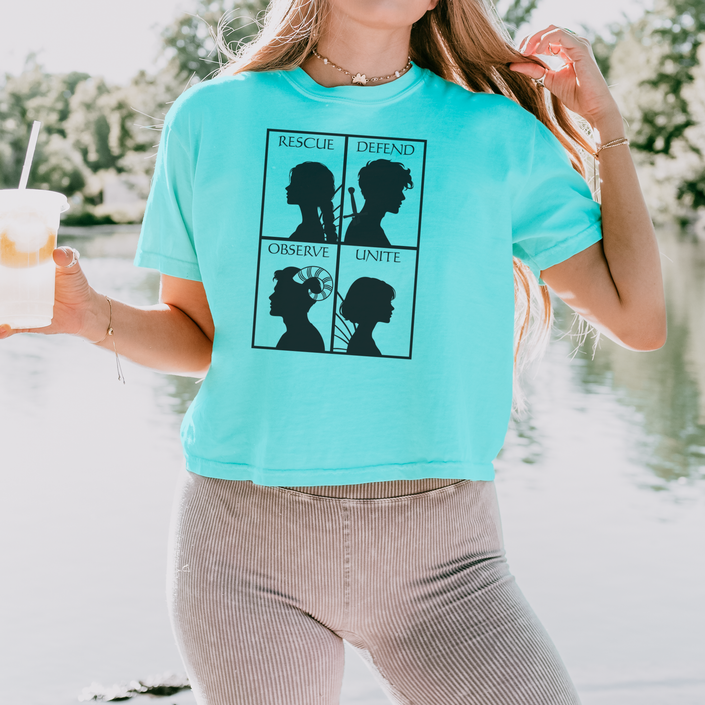 A woman wearing a bright turquoise graphic t-shirt with a black four-panel silhouette design, paired with beige corduroy pants, standing outdoors near water.