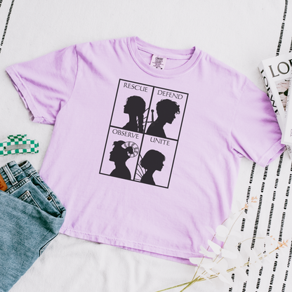 A light purple short-sleeve t-shirt with a black graphic print featuring silhouettes and the words 'Rescue', 'Defend', 'Observe', and 'Unite'. The shirt is styled in a flat-lay arrangement with blue jeans, a green and white checkered cloth, and a magazine on a white fabric background.