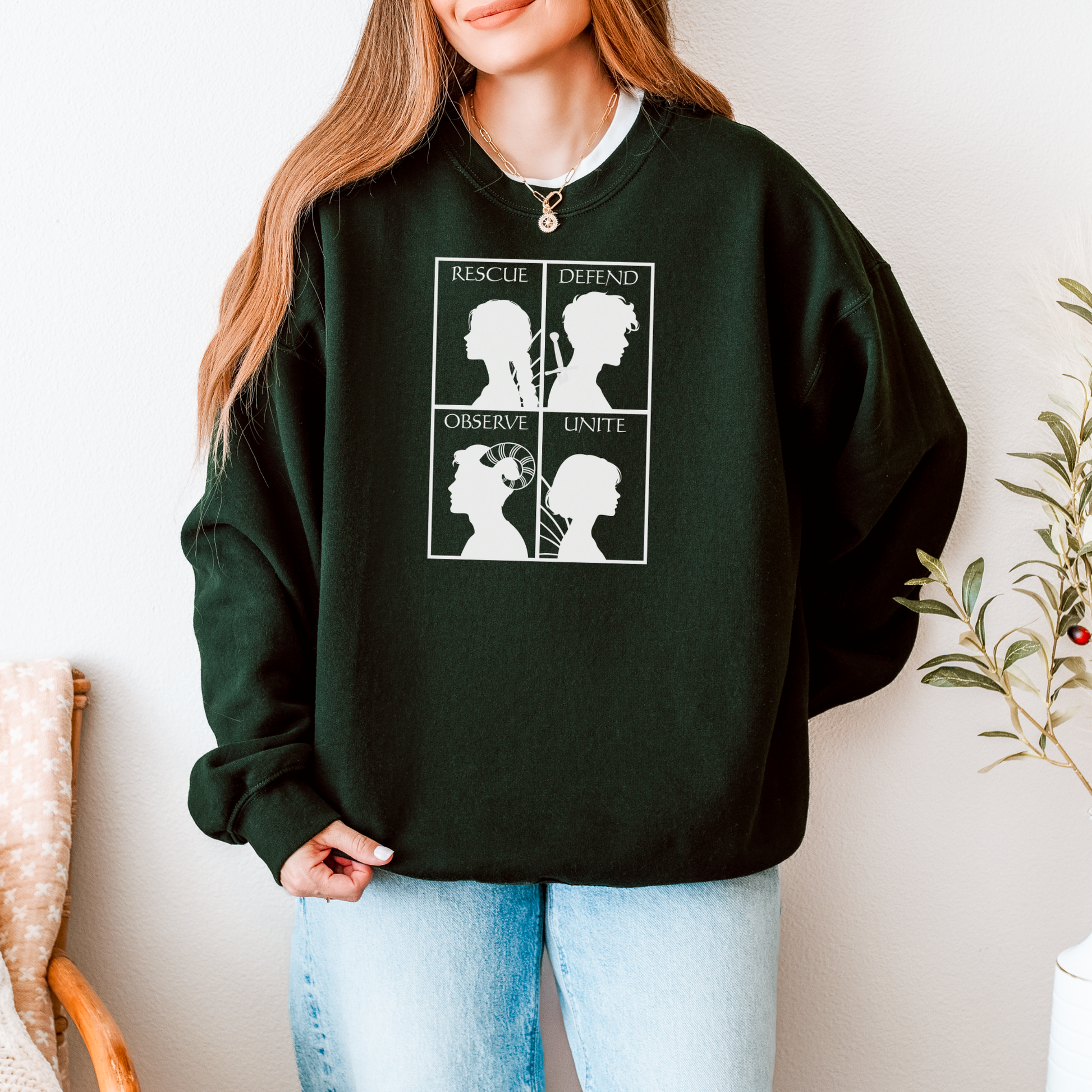 A woman wearing a dark forest green crewneck sweatshirt with a white graphic print featuring four quadrants and the words 'Rescue', 'Defend', 'Observe', and 'Unite'. She is paired with light blue jeans in a brightly lit indoor setting.