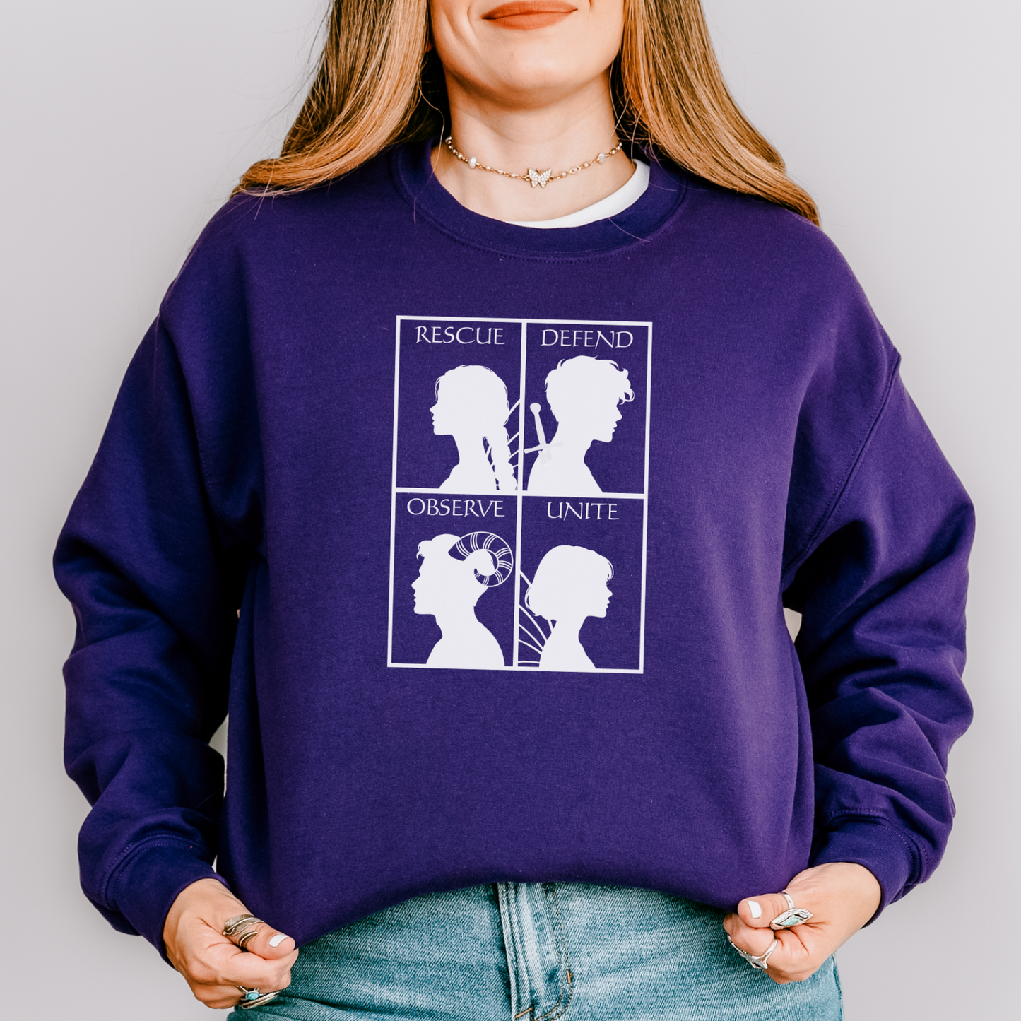 A woman wearing a purple crewneck sweatshirt with a white graphic print on the front, paired with light blue jeans, photographed against a neutral gray background.