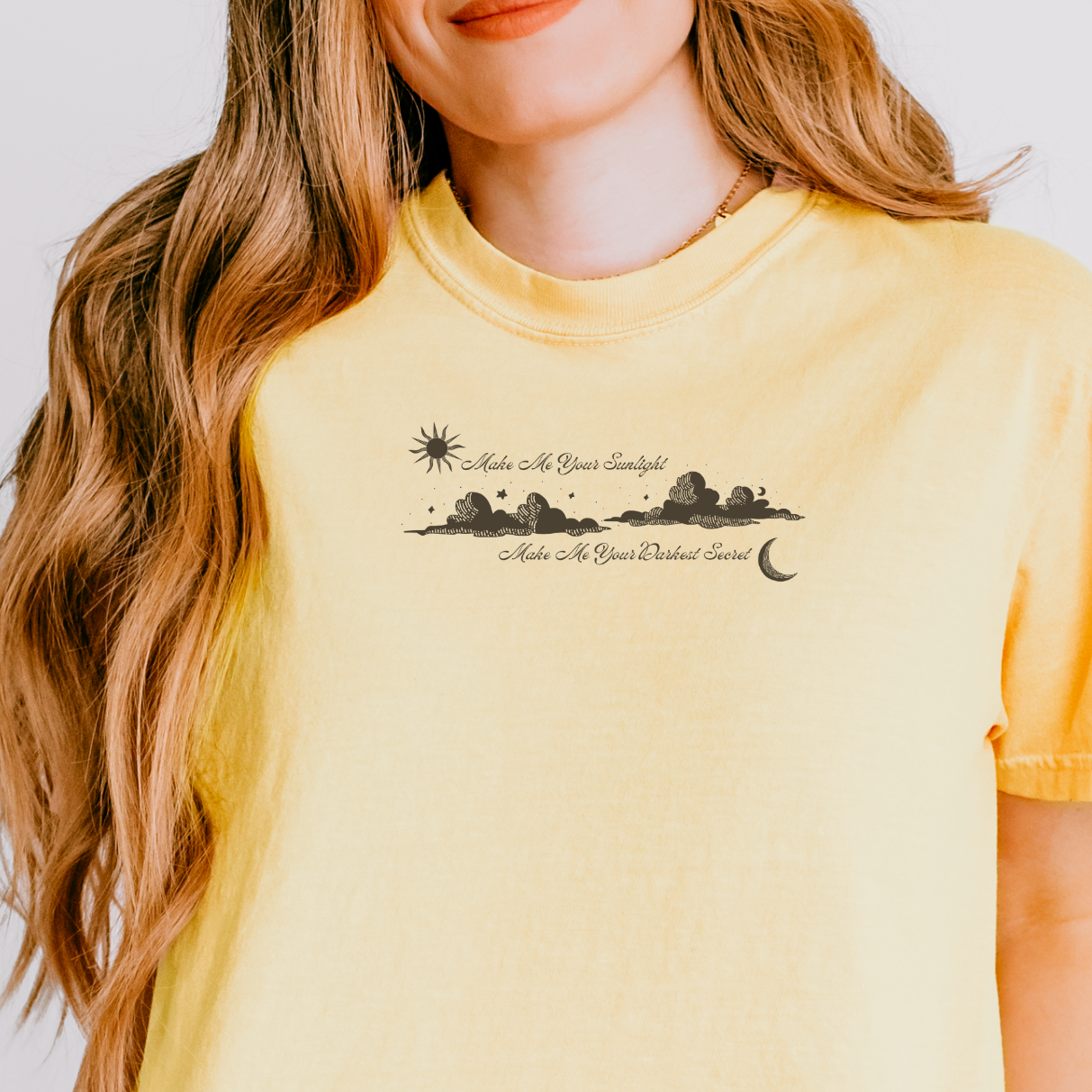 A woman wearing a pale yellow short-sleeve t-shirt with a black graphic print of a sun, mountains, and a crescent moon on the chest, paired with blue jeans against a light gray background.