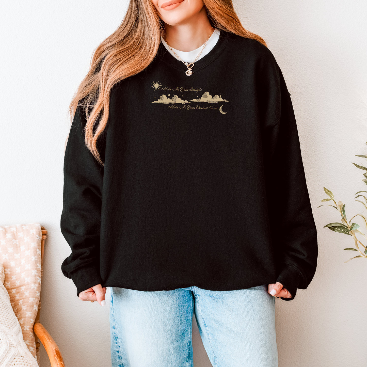 A woman wearing a black crewneck sweatshirt with a gold-toned desert landscape graphic, paired with light blue jeans. The shirt features the text 'Make Me Your Sunlight' and 'Make Me Your Darkest Secret'.