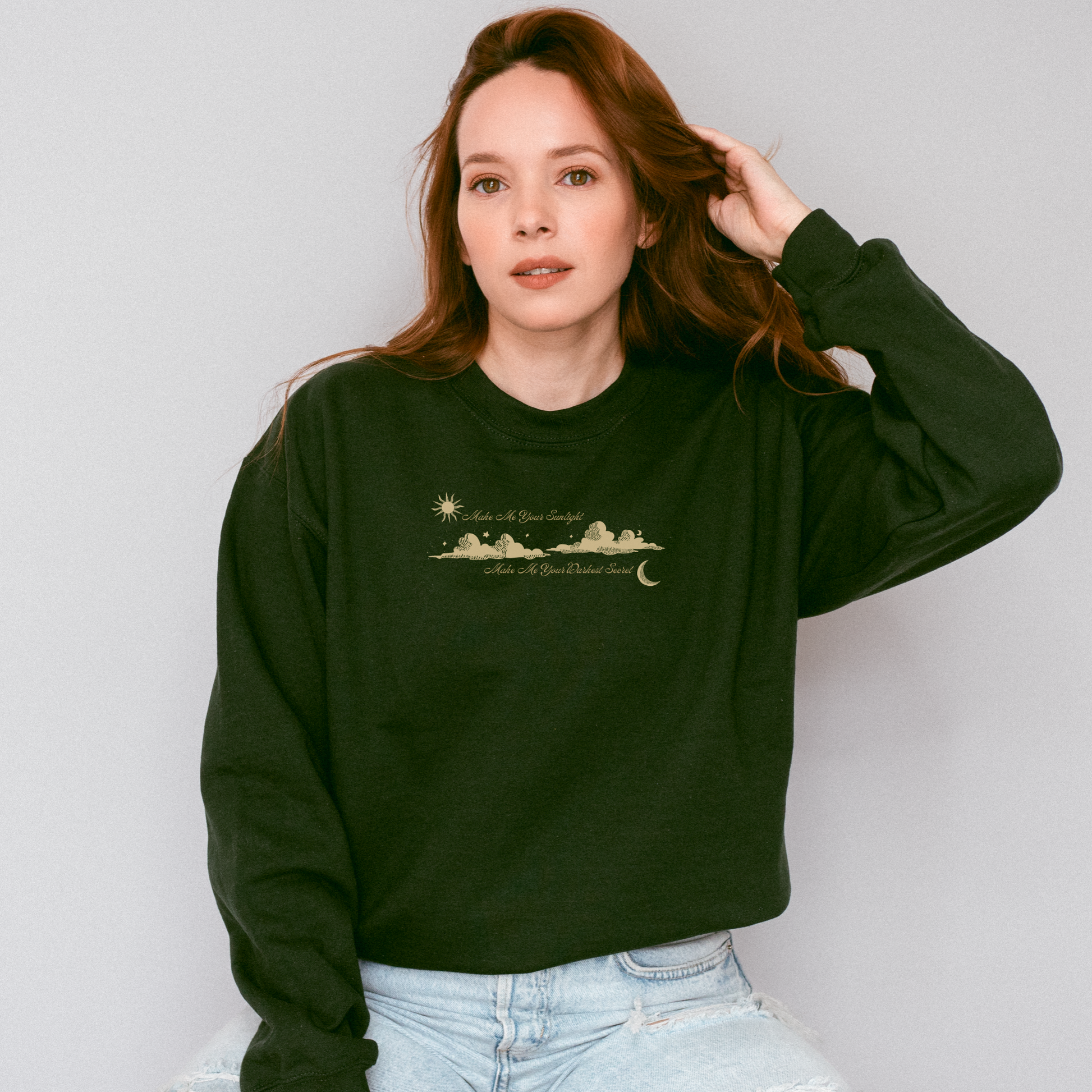 A woman with long red hair wearing a dark forest green crewneck sweatshirt with a gold-toned celestial landscape graphic on the chest, paired with light-wash jeans, set against a plain light gray background. The shirt features the text 'Make Me Your Sunlight' and 'Make Me Your Darkest Secret'.