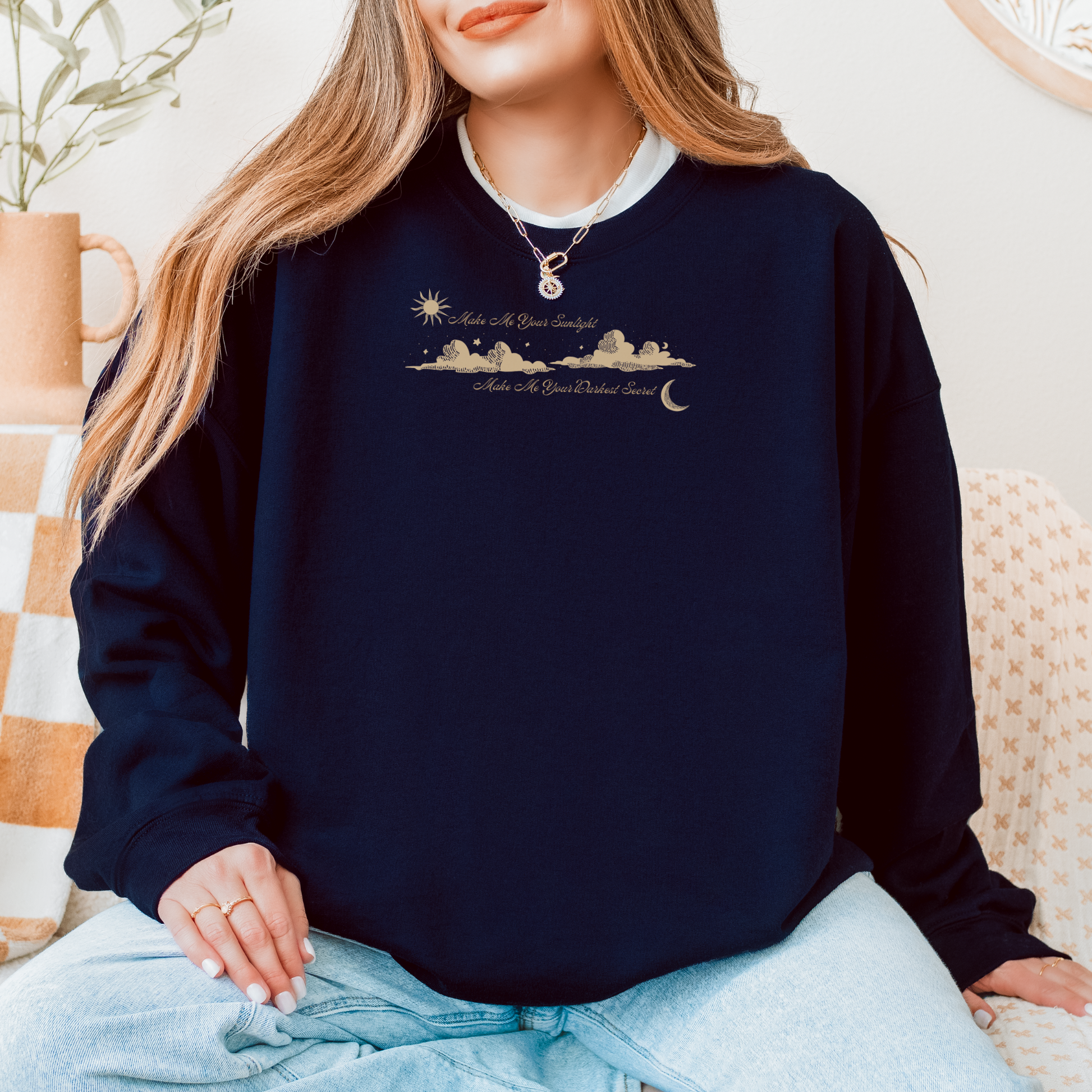 A woman wearing a navy blue crewneck sweatshirt with a gold celestial graphic and text on the chest, paired with light blue jeans in a cozy indoor setting. The shirt features the text 'Make Me Your Sunlight' and 'Make Me Your Darkest Secret'.