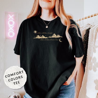 A woman wearing a black comfort-fit graphic t-shirt with a gold celestial design of a sun, moon, and stars. The shirt features the text 'Make Me Your Sunlight' and 'Make Me Your Darkest Secret'.