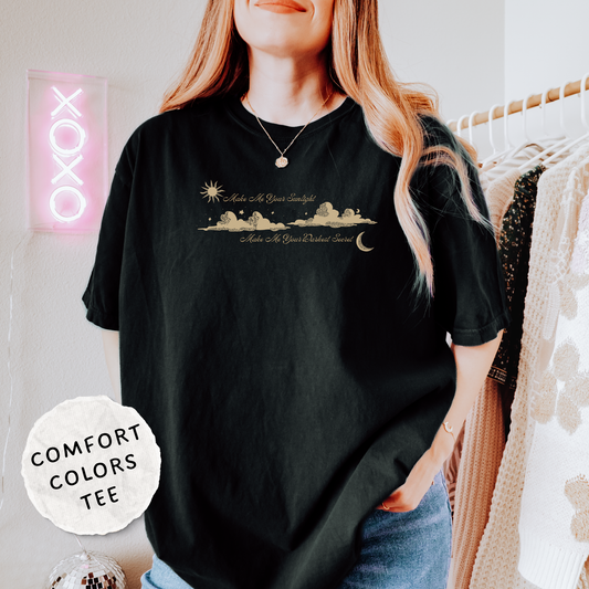 A woman wearing a black comfort-fit graphic t-shirt with a gold celestial design of a sun, moon, and stars. The shirt features the text 'Make Me Your Sunlight' and 'Make Me Your Darkest Secret'.