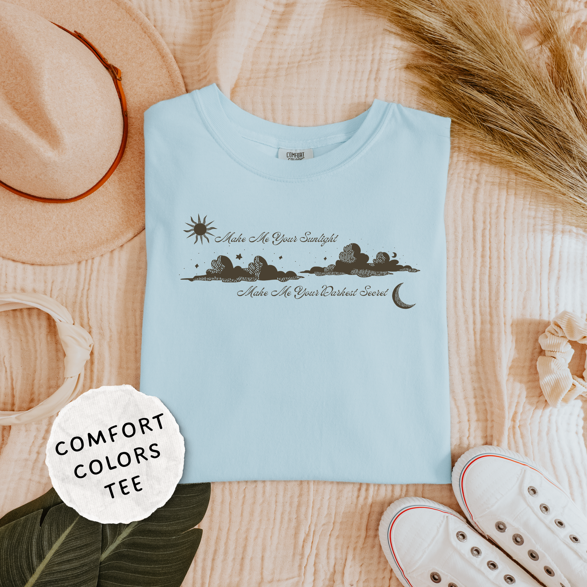 A light blue Comfort Colors t-shirt with a black graphic of a star, moon, and landscape, styled in a flat lay with a beige hat, white sneakers, dried grass, and a 'COMFORT COLORS TEE' label. The shirt features the text 'Make Me Your Sunlight' and 'Make Me Your Darkest Secret'.