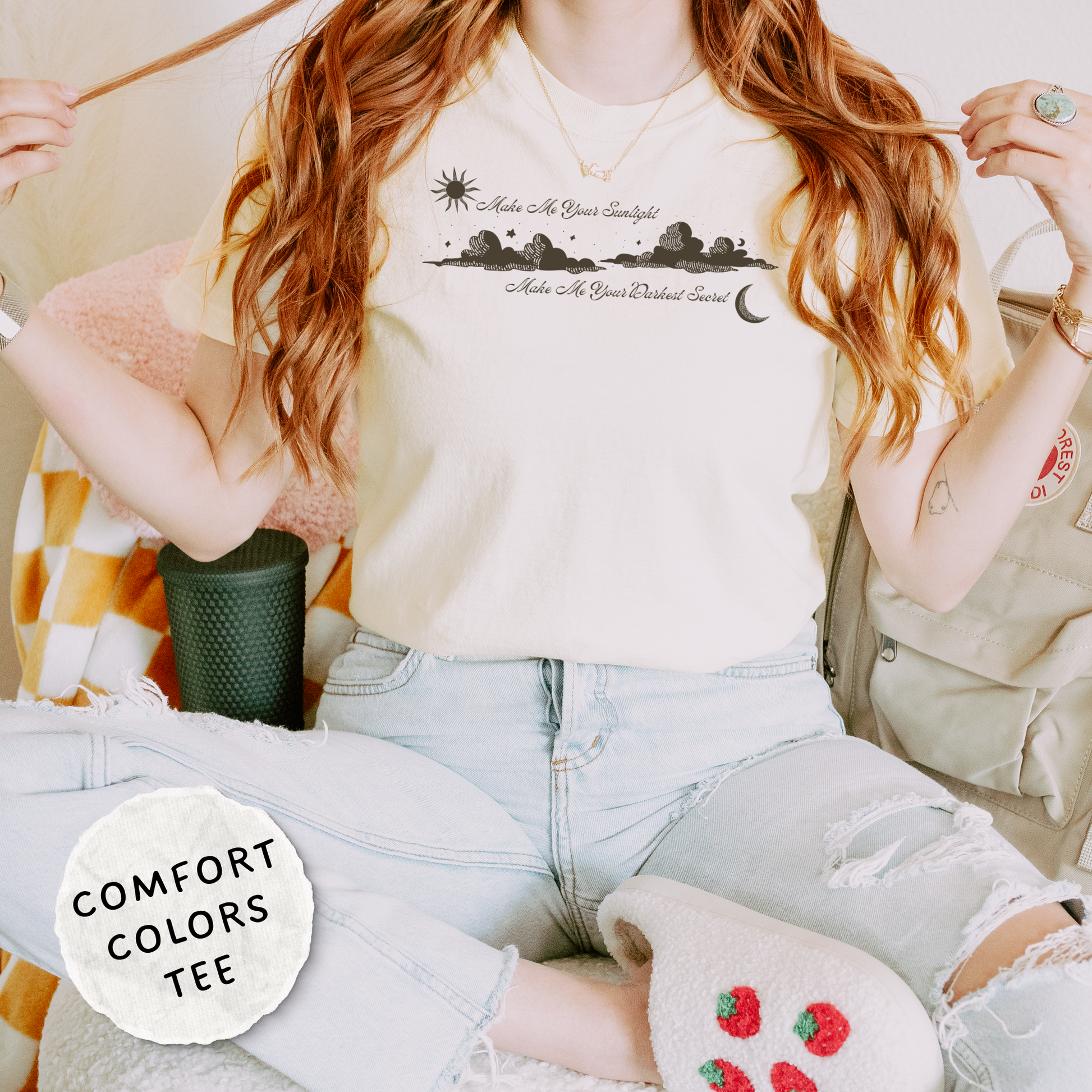 A woman wearing a Comfort Colors graphic t-shirt with a black sunset landscape print and text, paired with light-wash ripped jeans and cozy slippers, in a cozy indoor setting. The shirt features the text 'Make Me Your Sunlight' and 'Make Me Your Darkest Secret'.