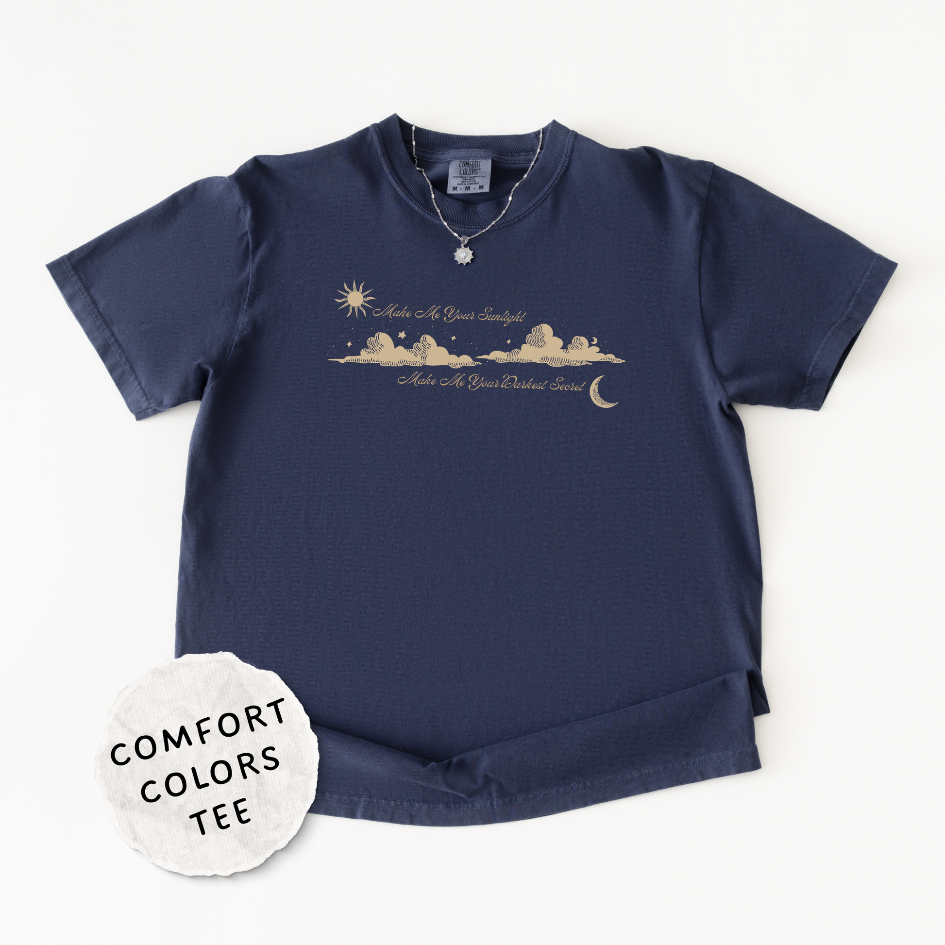 A navy blue short-sleeve t-shirt with a gold-toned graphic of a sunrise and sunset over mountains. The shirt features the text 'Make Me Your Sunlight' and 'Make Me Your Darkest Secret'. A silver necklace is draped over the collar.