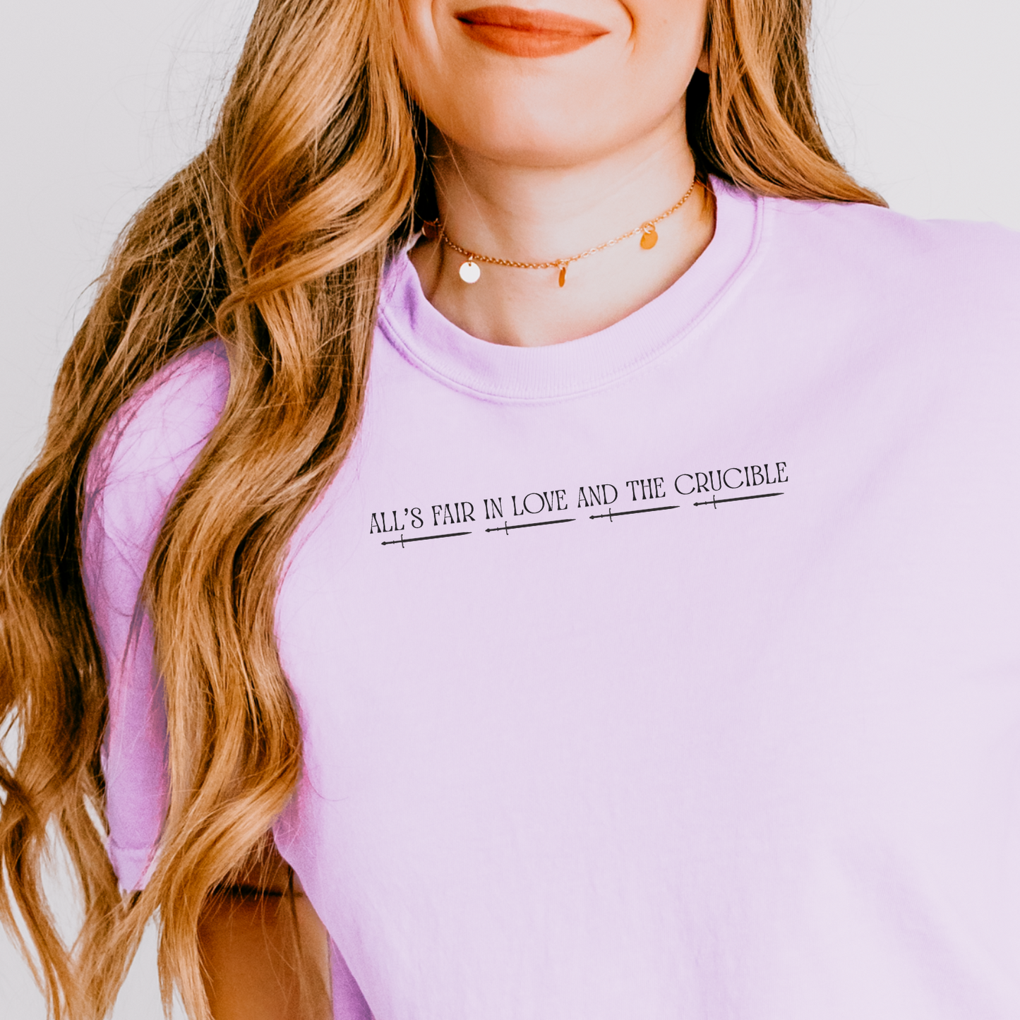 A woman wearing a light purple/lavender short-sleeve t-shirt with black text reading 'ALL'S FAME IN LOVE AND THE Cruicible.' on the chest, shown against a plain light background.