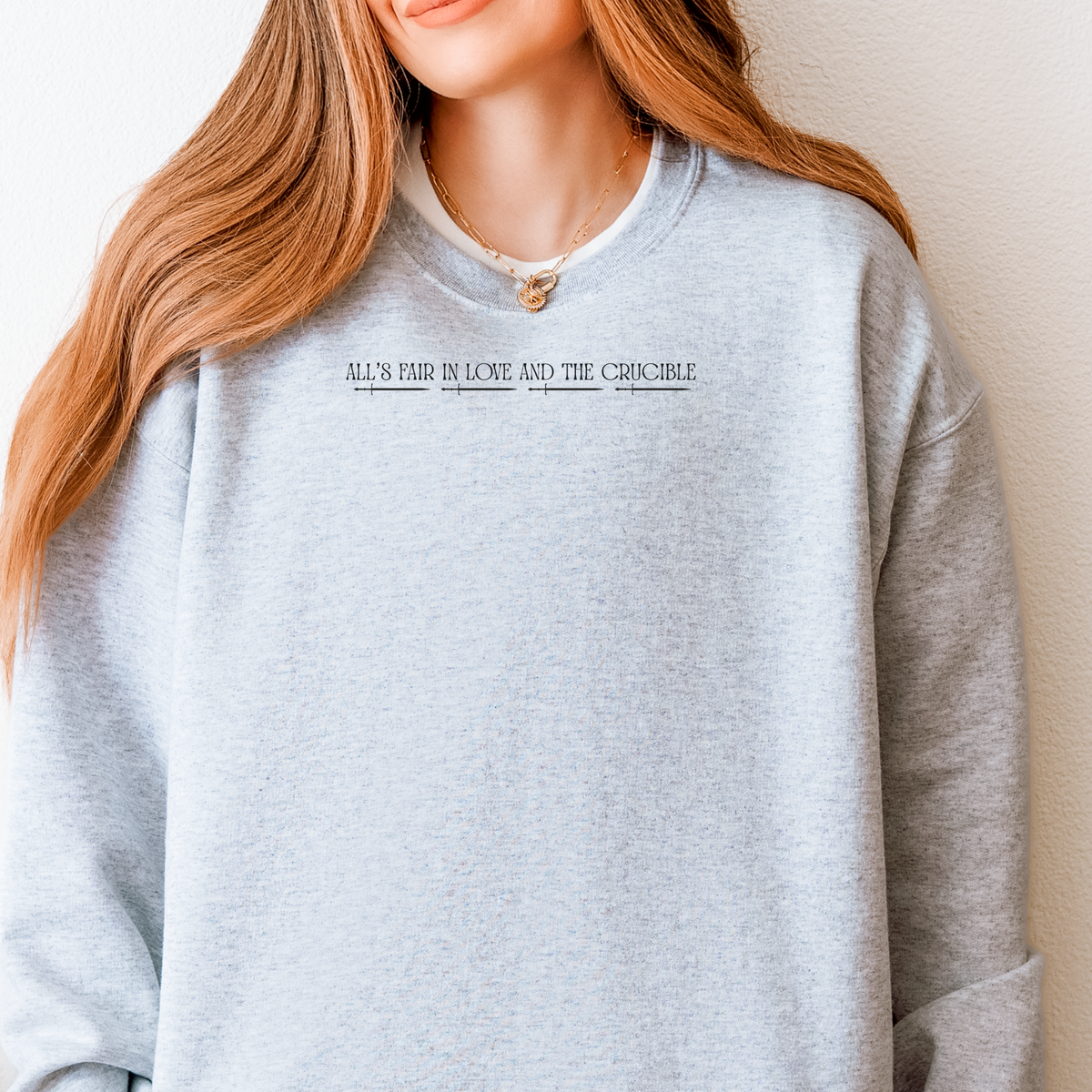 A woman wearing a light heather gray crewneck sweatshirt with the text 'ALL'S FAIR IN LOVE AND THE CRUCIBLE' printed in black across the chest.