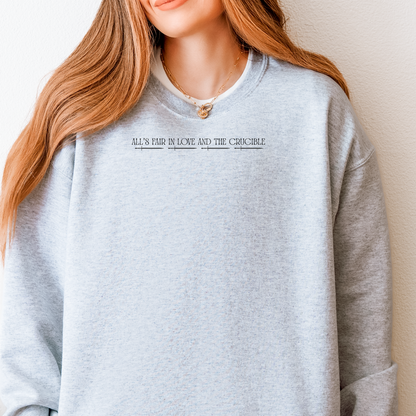 A woman wearing a light heather gray crewneck sweatshirt with the text 'ALL'S FAIR IN LOVE AND THE CRUCIBLE' printed in black across the chest.
