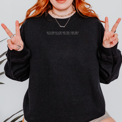 A person wearing a black crewneck sweatshirt with white text across the chest reading 'ALL'S FAIR IN LOVE AND THE CRUCIBLE'. The person has red hair, is wearing layered gold necklaces, and is posing with peace signs.