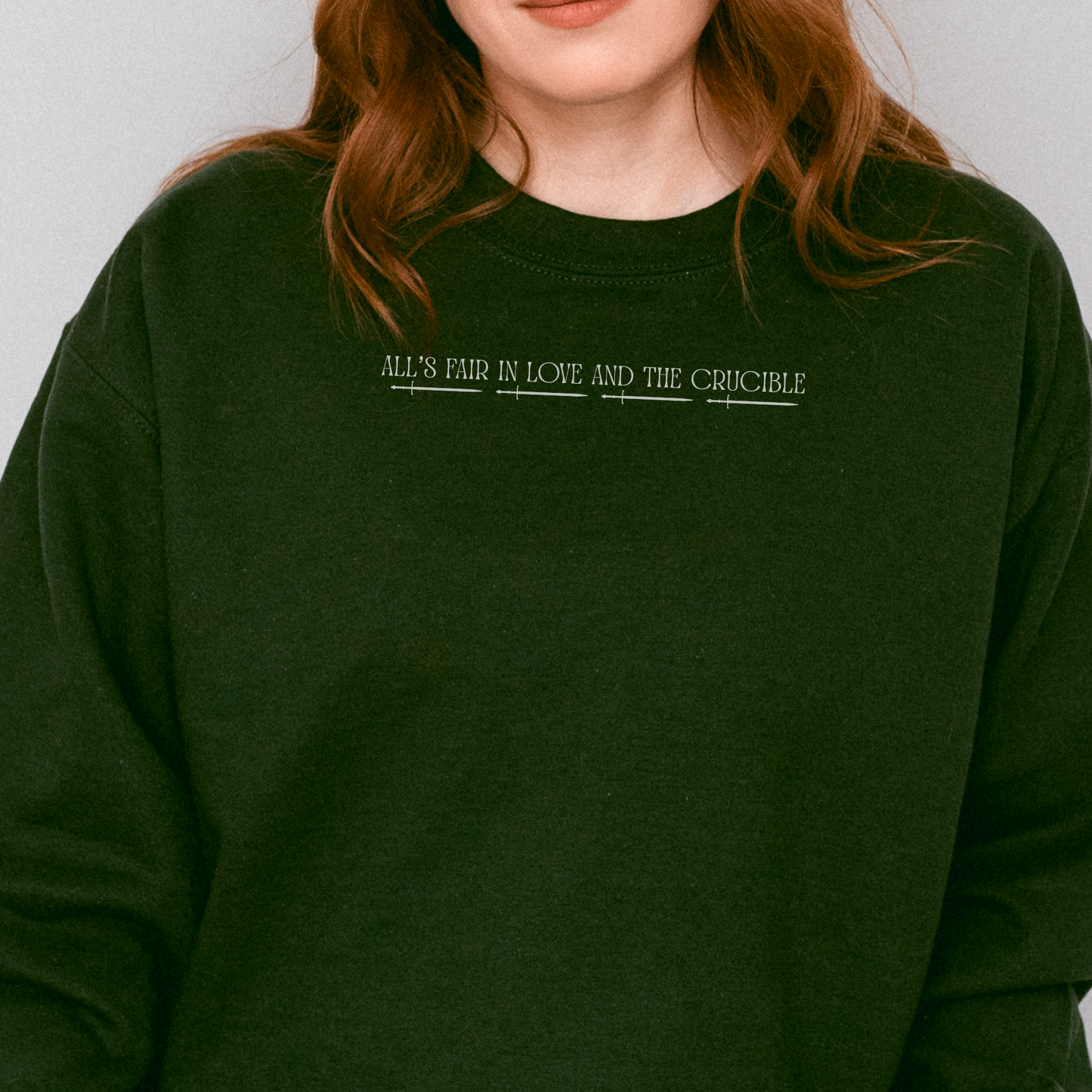 A close-up of a person wearing a dark forest green crewneck sweatshirt with the text 'ALL'S FAIR, in LOVE AND THE CRUCIBLE' printed in white across the chest.