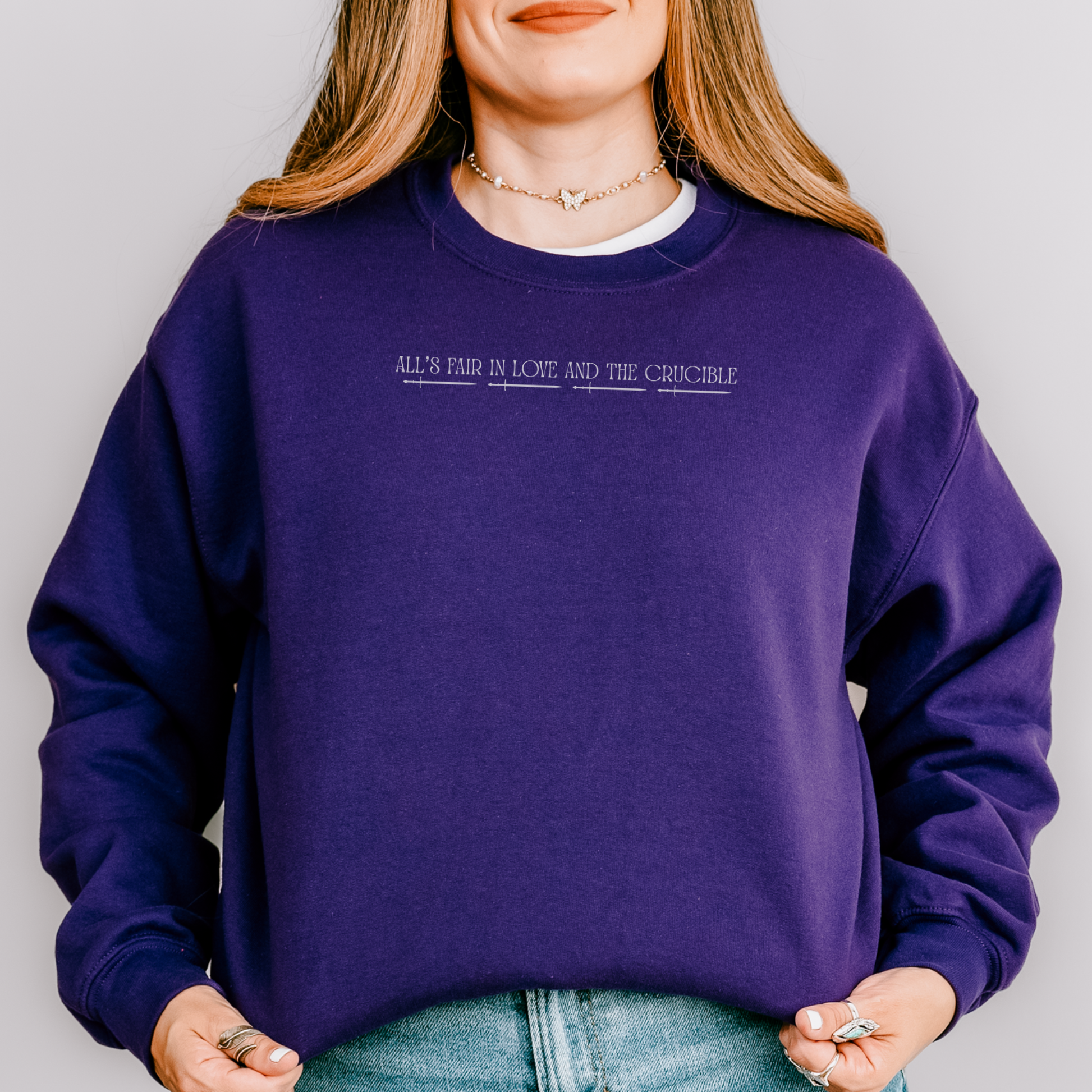 A woman wearing a dark purple crewneck sweatshirt with the text 'ALL'S FAIR IN LOVE AND THE CRUCIBLE.' printed in white across the chest, paired with light blue jeans against a solid gray background.