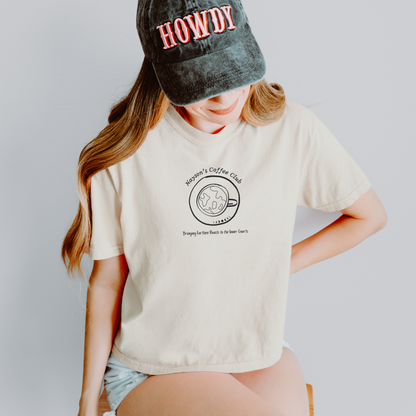 A woman wearing a cream-colored graphic t-shirt with a black coffee cup illustration and the words 'Nayson's Coffee Club'. She is also wearing a dark gray washed baseball cap with 'HOWDY' in red text.