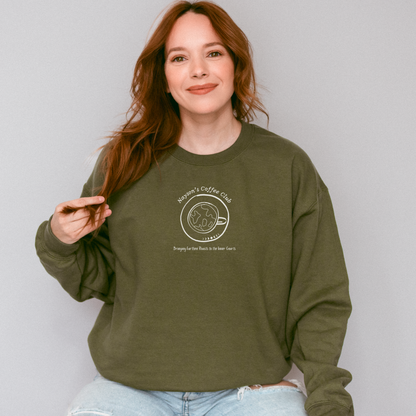 A woman with long red hair wearing an olive green crewneck sweatshirt reads 'Nayson's Coffee Club' and 'Bringing Earthen Roasts to the Inner Courts'. She is wearing light-wash blue jeans against a plain light gray studio background.