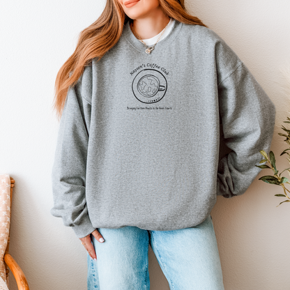 A woman wearing a heather gray crewneck sweatshirt with a black coffee-themed graphic on the chest that reads 'Nayson's Coffee Club' and 'Bringing Earthen Roasts to the Inner Courts', paired with light blue jeans, standing against a neutral wall.