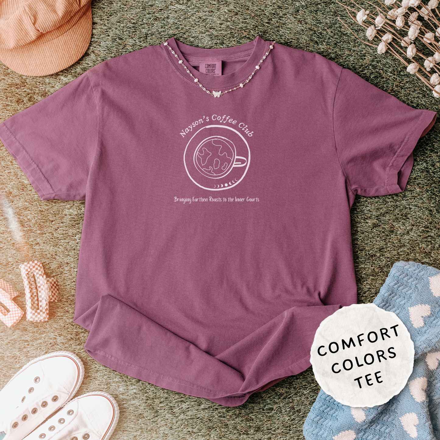A mauve purple short-sleeve t-shirt with a white graphic reading 'Noonan's Coffee Club' and a coffee cup/donut illustration, styled in a flat lay on a green textured surface with a peach cap, white shoes, a necklace, and a blue heart-patterned blanket nearby. A white circular badge says 'COMFORT COLORS TEE'.