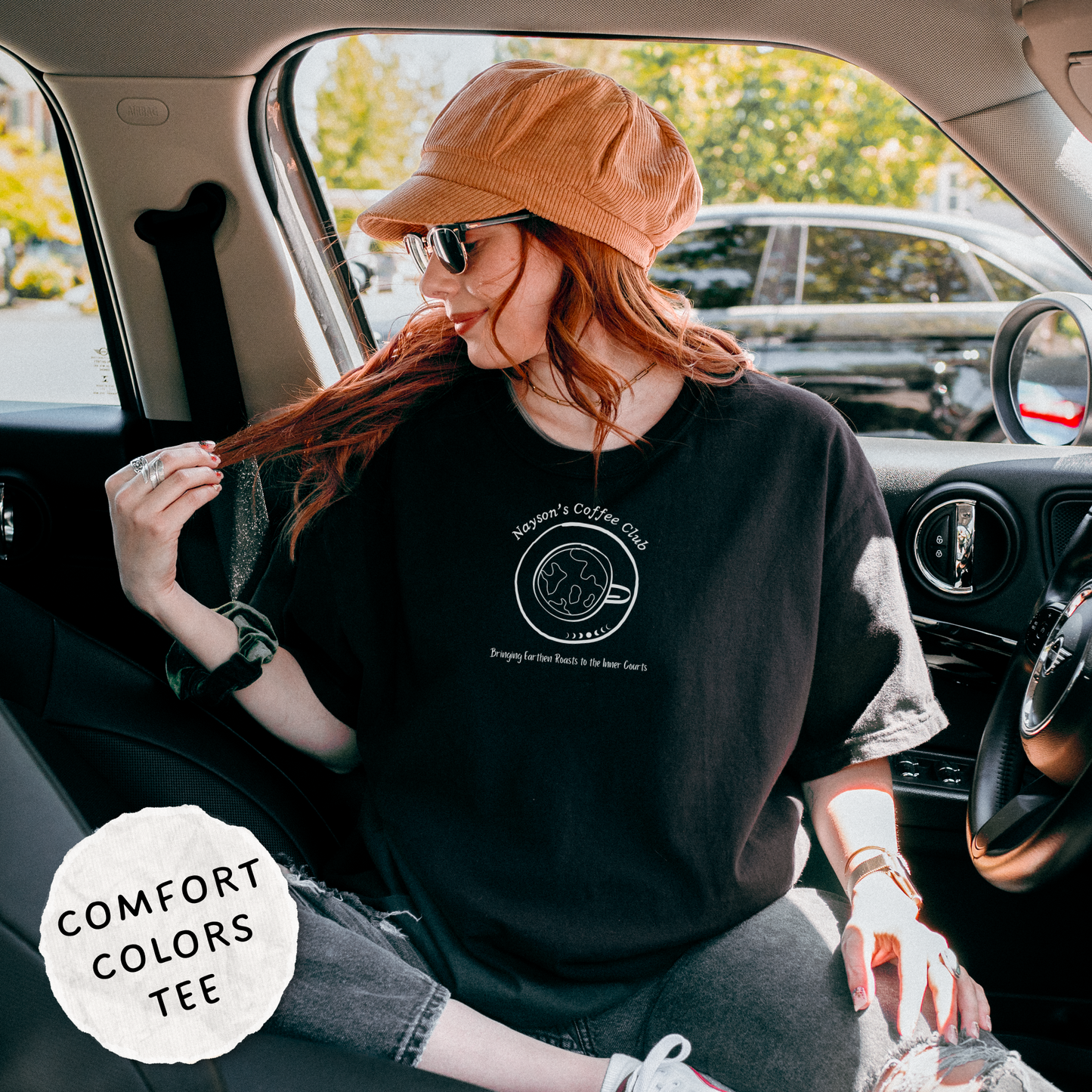 A woman sitting in the driver's seat of a car wearing a black Comfort Colors graphic t-shirt, a brown corduroy cap, and sunglasses. The t-shirt features a white circular logo that reads 'Comfort Colors Coffee Club'.