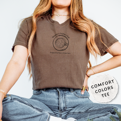 A woman wearing a brown v-neck t-shirt with a black coffee cup graphic and text, paired with blue jeans. A white circular badge on the right reads 'COMFORT COLORS TEE'.