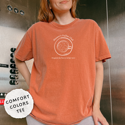 A woman wearing an oversized washed orange t-shirt with a white graphic that reads 'Nayson's Coffee Club'. She is standing in front of a metallic elevator wall, with a 'COMFORT COLORS TEE' badge overlay in the bottom left corner.
