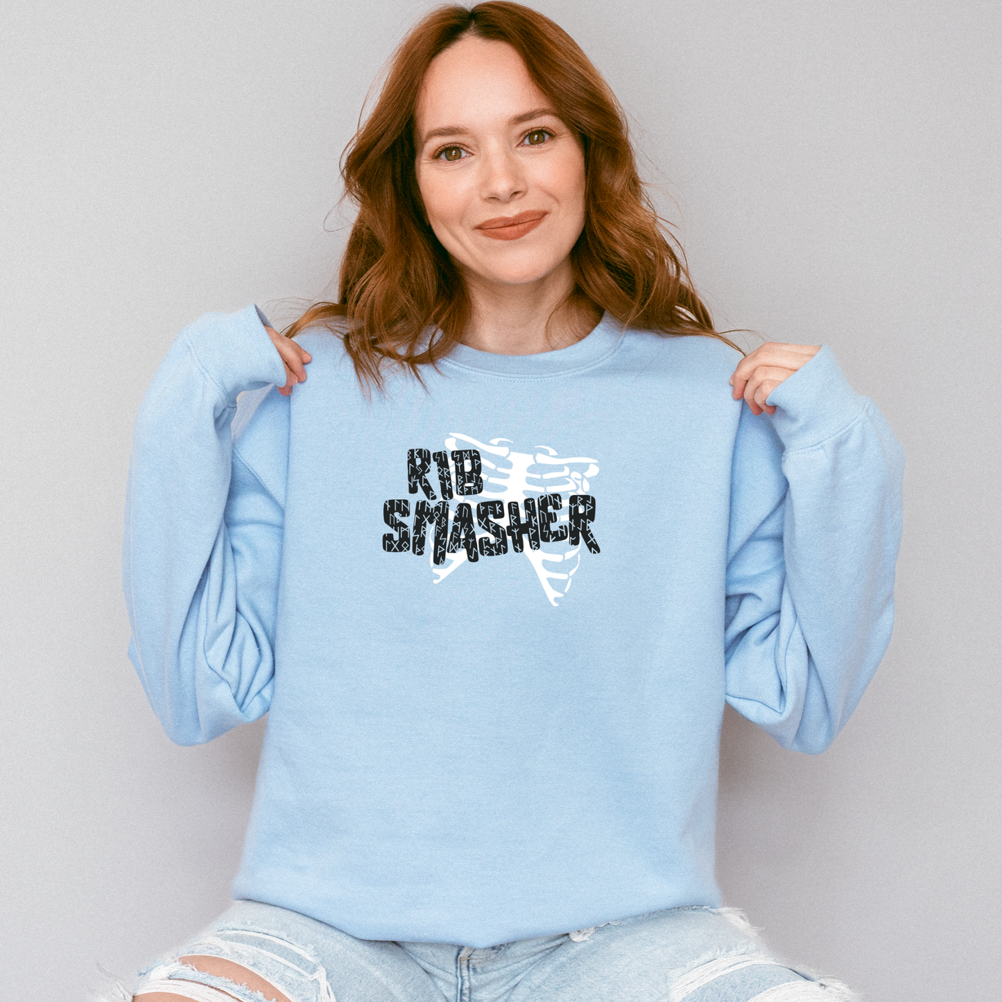 Woman wearing a light blue sweatshirt with 'KID SMASHER' text, sitting against a plain background.