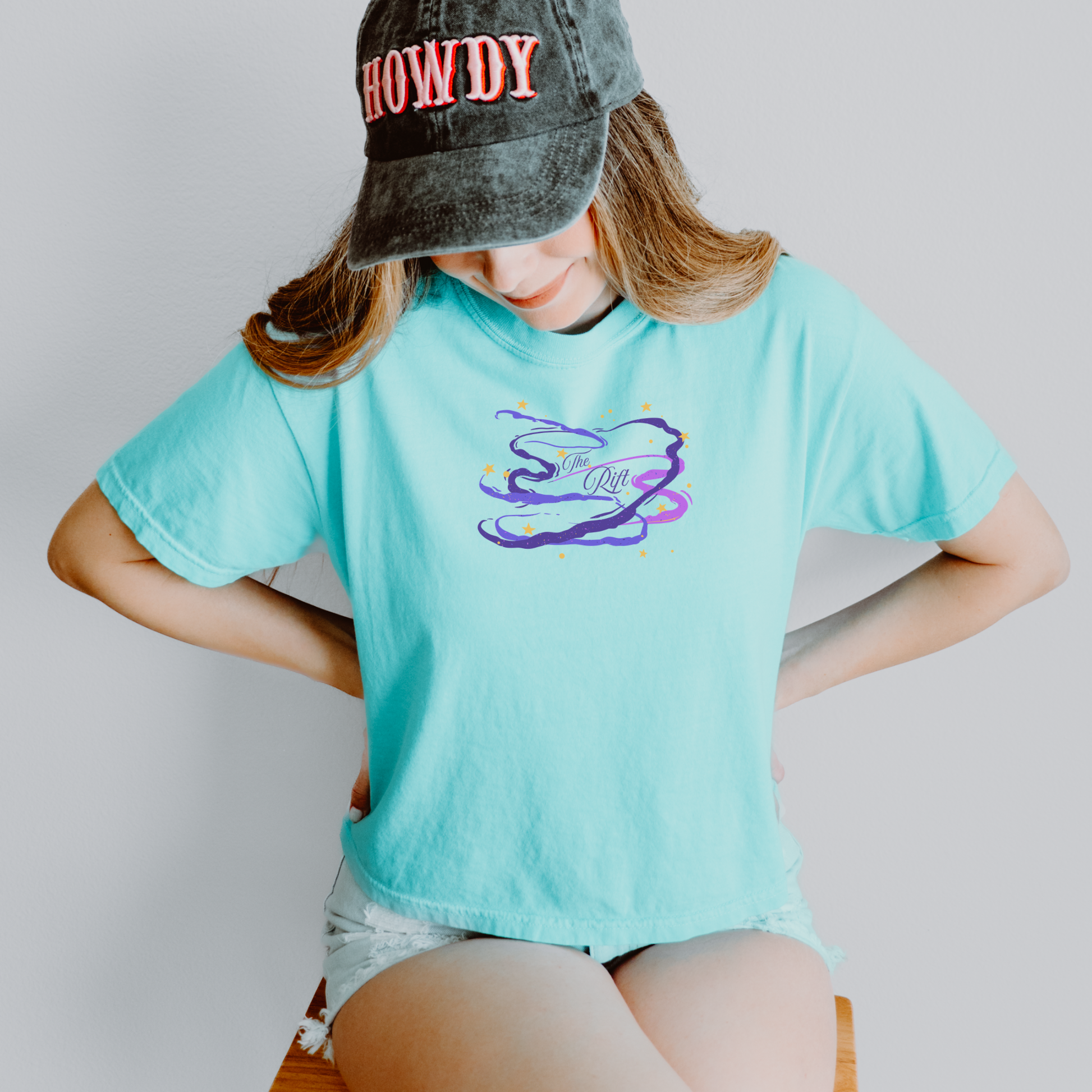 A woman wearing a light aqua blue graphic t-shirt with a purple and blue swirl design on the chest, paired with a dark gray acid-wash baseball cap and denim shorts, sitting against a light gray background.