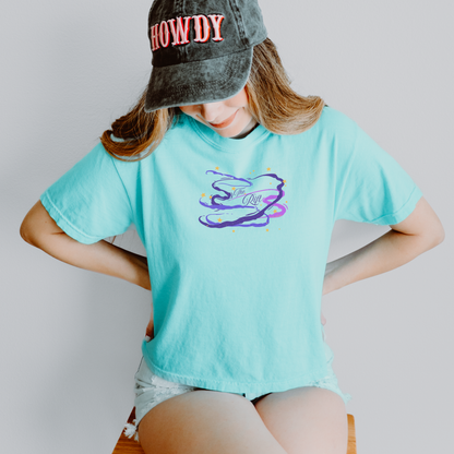 A woman wearing a light aqua blue graphic t-shirt with a purple and blue swirl design on the chest, paired with a dark gray acid-wash baseball cap and denim shorts, sitting against a light gray background.