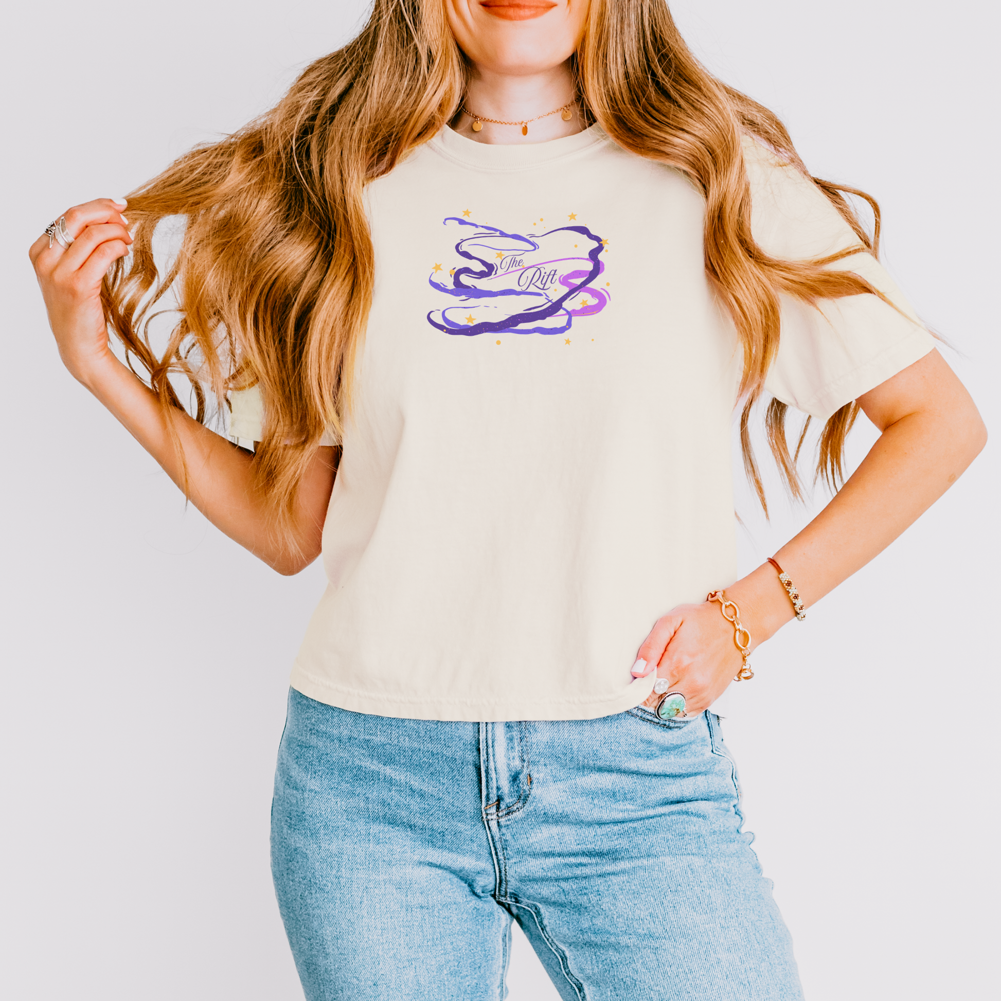 A woman wearing a cream-colored t-shirt with a purple and gold graphic of a stylized letter 'S' and the words 'Soul of a Girl', paired with light blue jeans, against a light gray background.