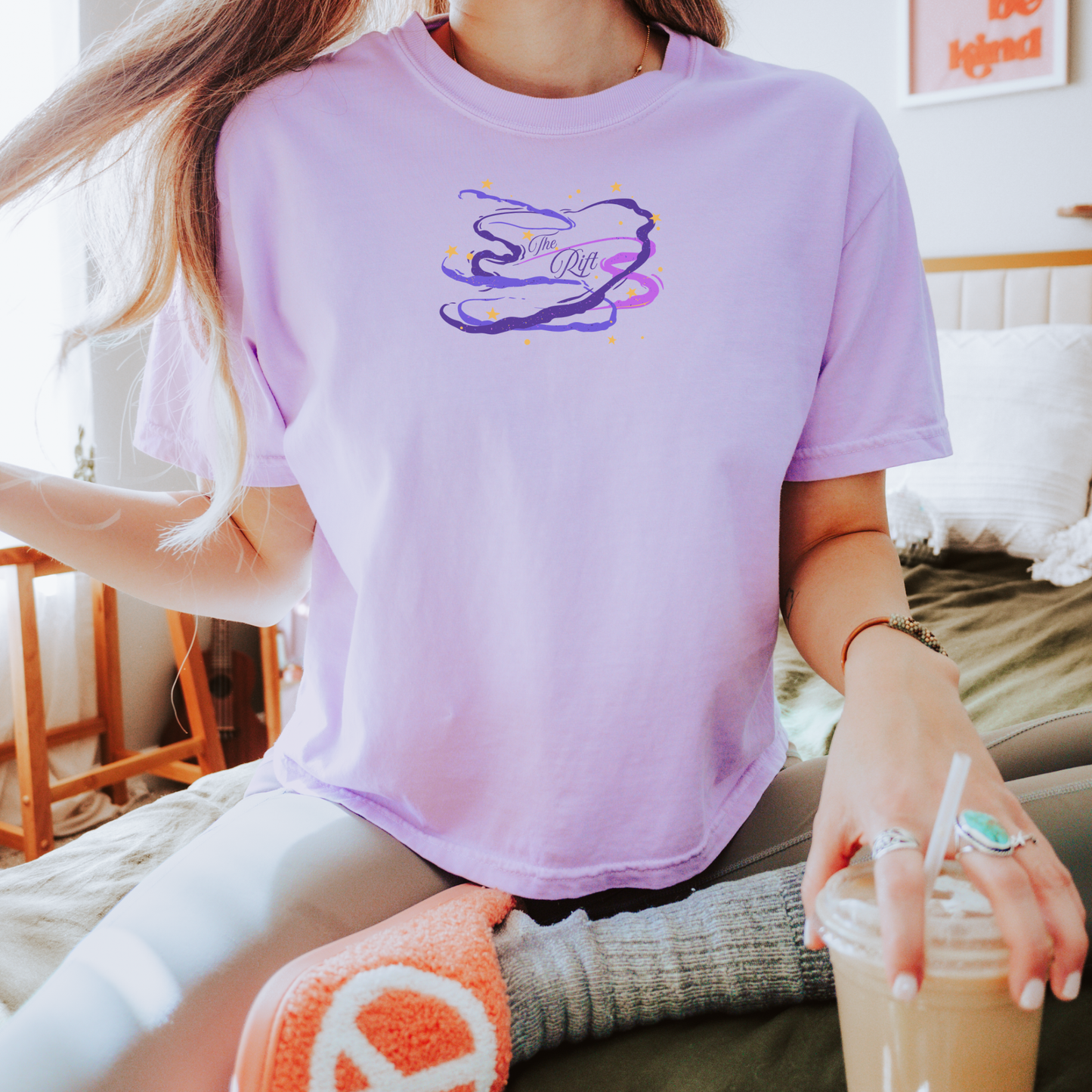 A person wearing a light lavender short-sleeve graphic T-shirt with a purple and yellow abstract cloud design on the chest, sitting in a bedroom setting and holding a drink.