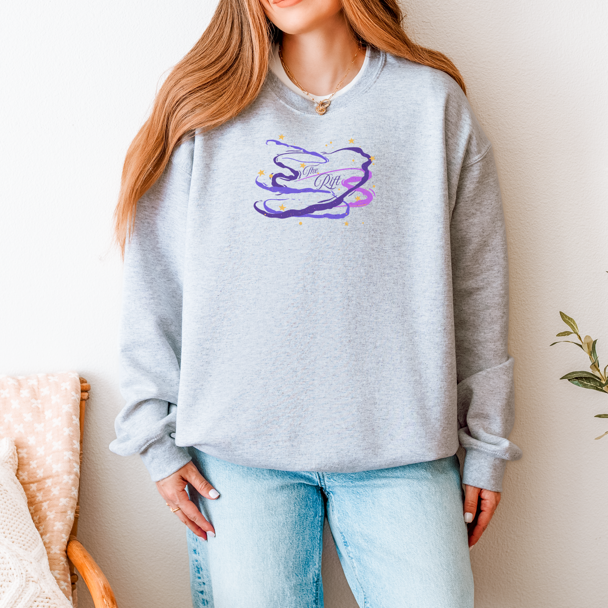 A woman wearing a light heather gray crewneck sweatshirt with a purple and blue abstract graphic on the chest, paired with light blue jeans.