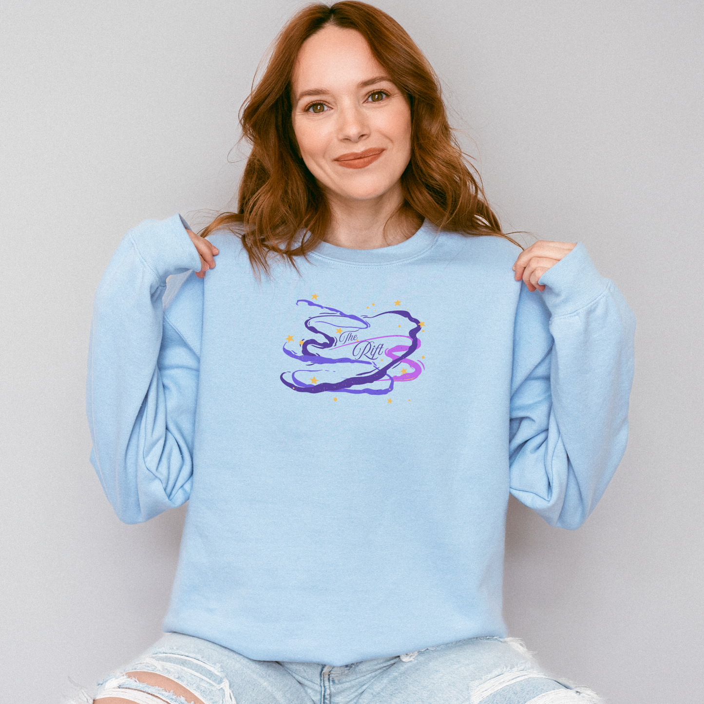 A woman with wavy light brown hair wearing a light blue crewneck sweatshirt with a colorful graphic on the chest, posing against a solid light gray background.