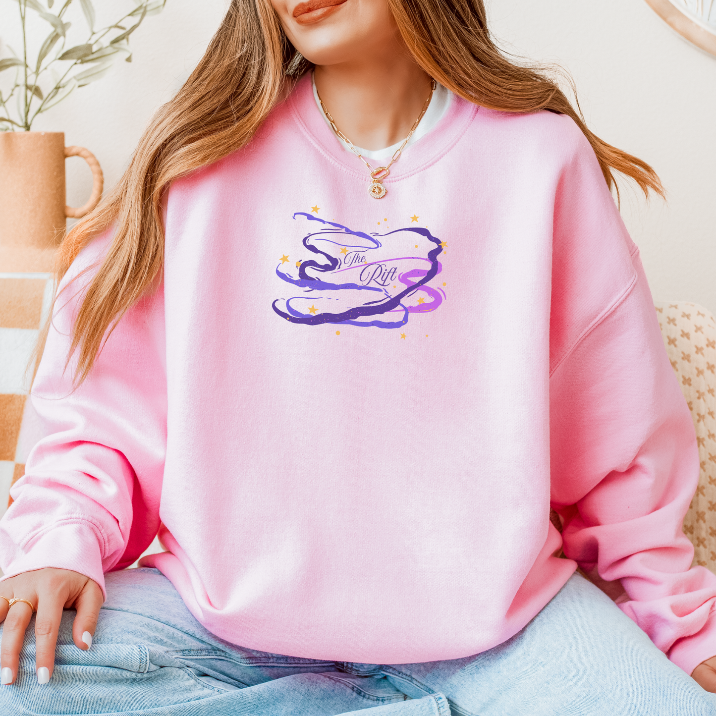 A woman wearing a pink crewneck sweatshirt with a purple and blue abstract cloud graphic on the chest, paired with light blue jeans in a cozy indoor setting.