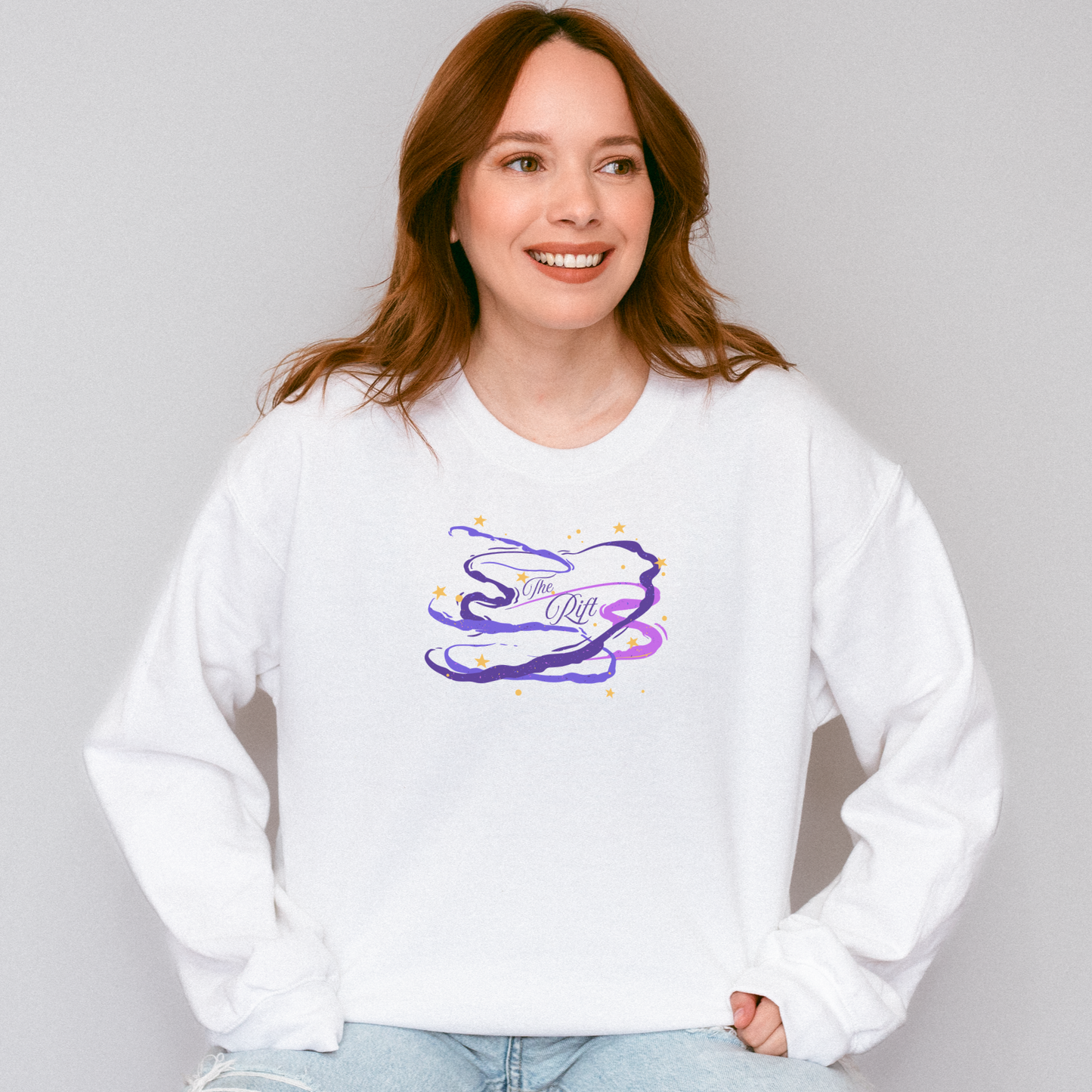 A woman with red hair wearing a white crewneck sweatshirt with a purple and blue abstract graphic on the chest, paired with light blue jeans, against a plain light gray background.