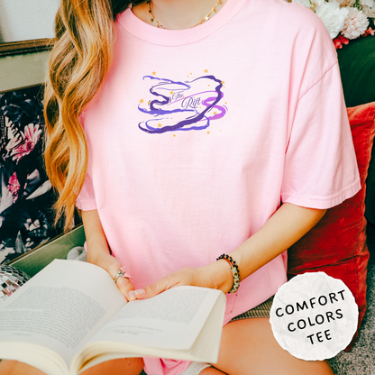 A woman wearing a light pink oversized T-shirt with a purple and white graphic on the chest, holding an open book while sitting on a red cushion. A white circular badge in the bottom right reads 'COMFORT COLORS TEE'.